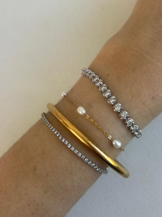 A close-up of a person's wrist wearing three delicate bracelets: a thick gold bangle, a thin gold chain with white pearls, and two silver-toned tennis bracelets with clear stones. By Nicola Coco & Lola 
