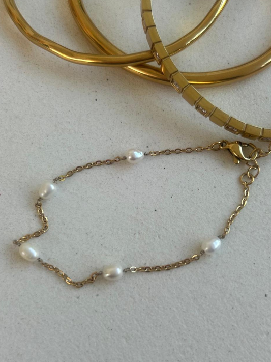 By Nicola delicate gold chain bracelet with five small white freshwater pearls spaced along the chain, displayed on a light textured surface with gold bangles in the background.