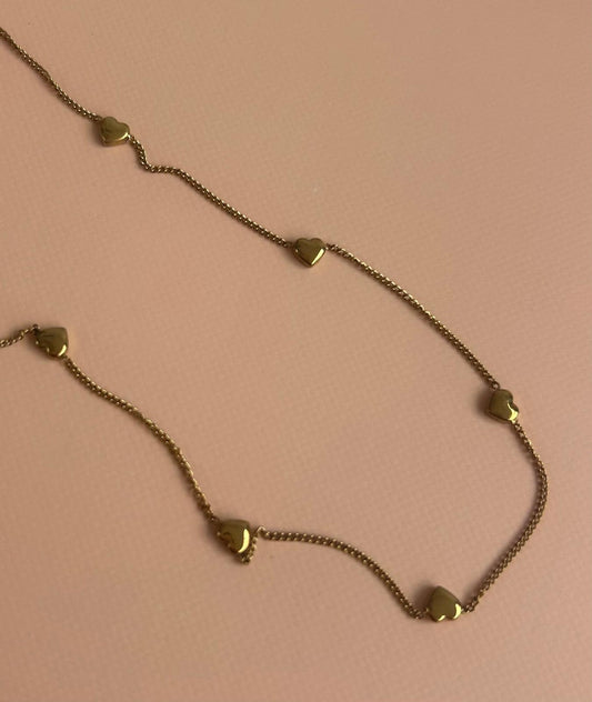 A gold chain necklace featuring five small, polished heart charms spaced along the chain, displayed on a solid light pink background. Exclusive to Coco & Lola and By Nicola