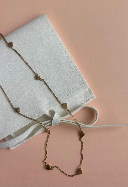 A delicate gold chain necklace with small heart charms, draped over a white fabric pouch on a soft peach-pink background. Exclusive to Coco & Lola and By Nicola