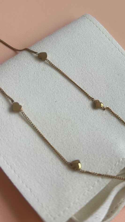 A delicate gold chain necklace with small, flat heart-shaped charms, displayed on a textured white fabric surface against a soft peach background. Exclusive to Coco & Lola and By Nicola